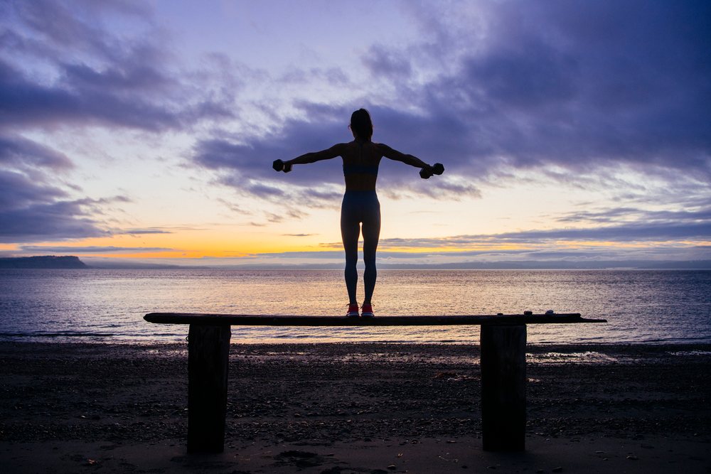Woman silhouetted at sunrise balances on a wooden beam at the beach while holding dumbbells, WA, USA.
