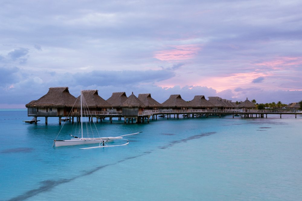Overwater bungalows and a sailboat at Bora Nui Resort during sunset beneath storm clouds, Tahiti, French Polynesia.