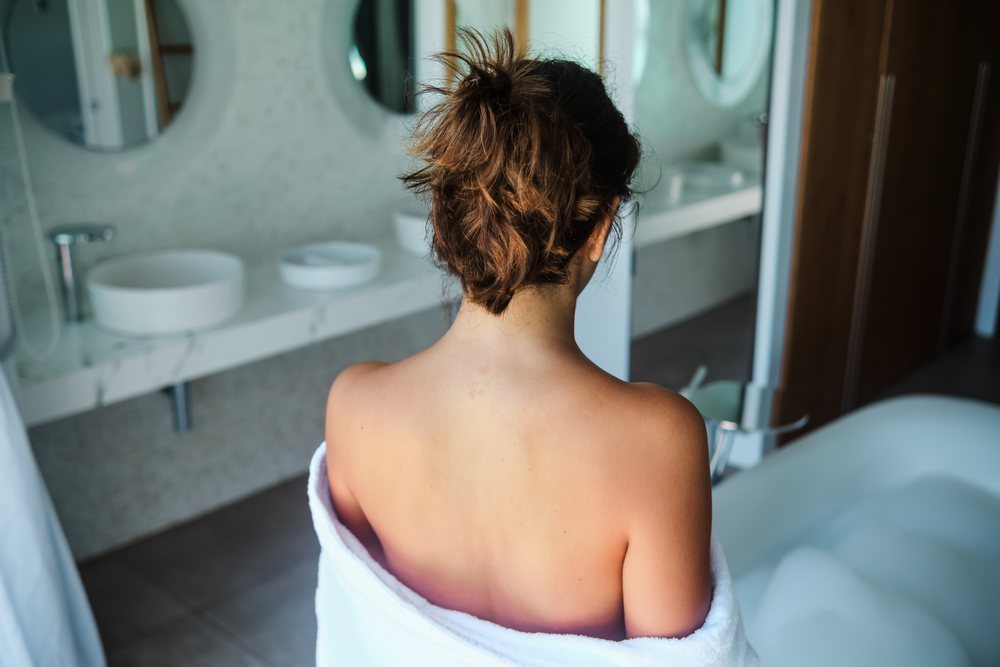 A woman wrapped in a towel is seen from behind sitting near a foam-filled bathtub in a modern bathroom, Maldives.