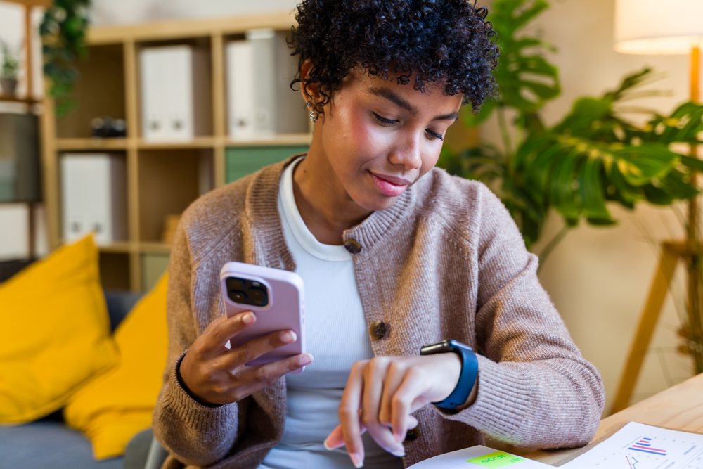 Woman sitting indoors at a table holding a smartphone and checking a smartwatch in a modern office setting with decorative plants and yellow pillows, Spain.
