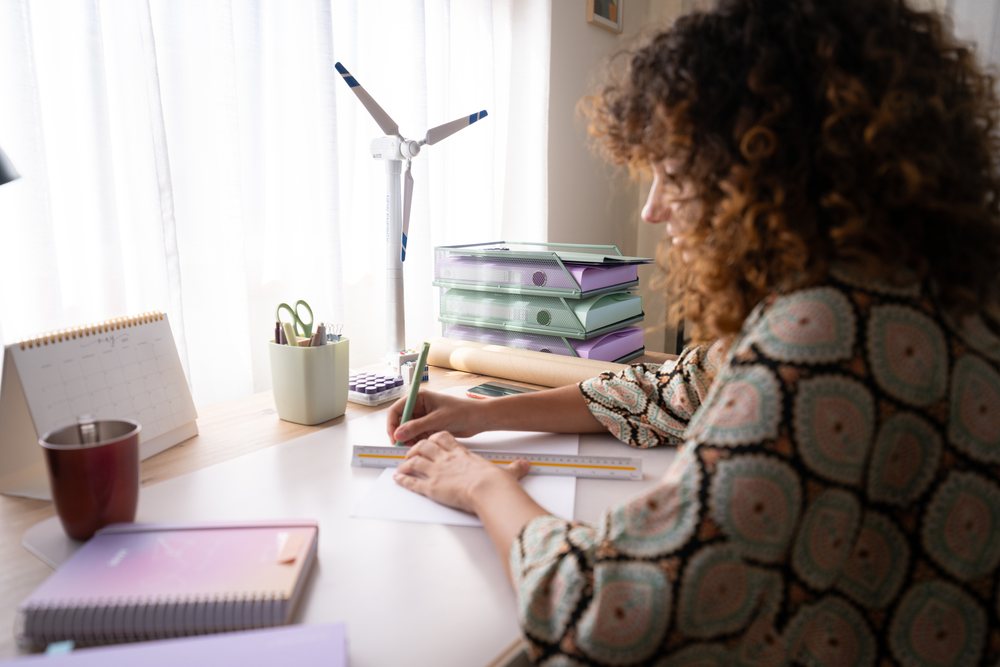 Woman with curly hair writes and measures paper using a ruler at an organized desk with stationery and a model wind turbine in a bright room, home, girl, Spain.