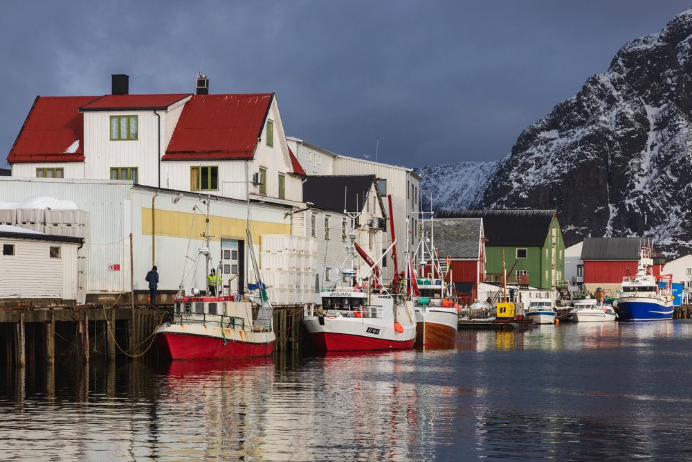 Fishing boats moored at a wooden dock in the harbor of Henningsvaer, with snow-covered mountains in the background and cloudy sky above, Landscape, Lofoten, Norway.