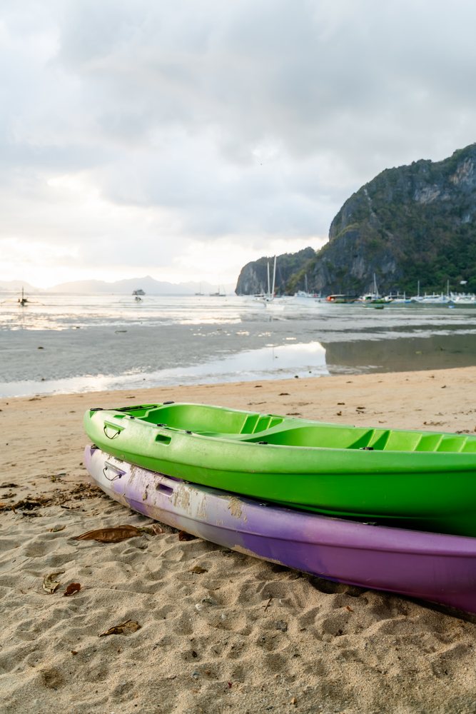 Two kayaks stacked on a sandy beach with boats floating in a bay, Philippines.