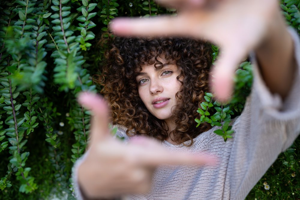 Woman with curly hair framing her face with hands outdoors in front of green leafy plants, home story, influencer, Spain.