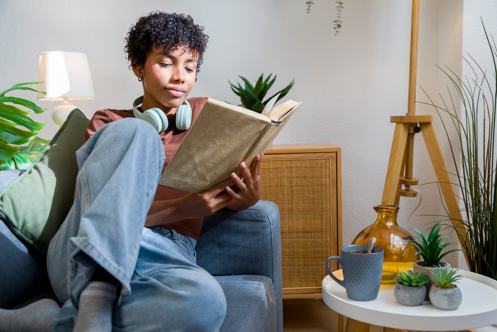 Young woman sits on a sofa reading a book with headphones around her neck and a coffee mug on a table in a cozy, daylight home setting.