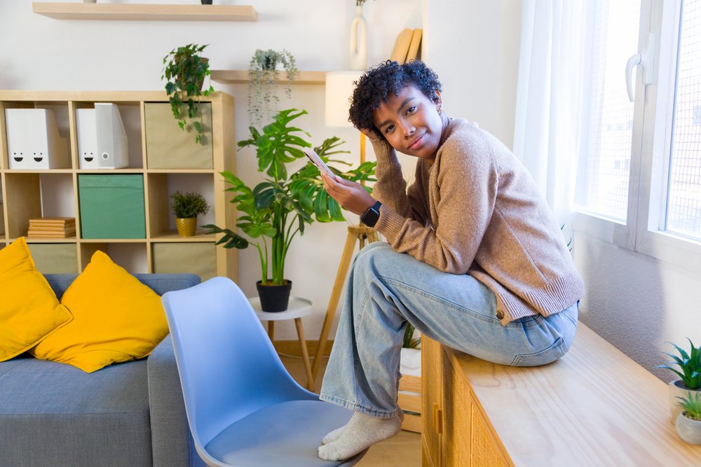 Young woman sits on a wooden cabinet at home near a window holding a phone with natural light.