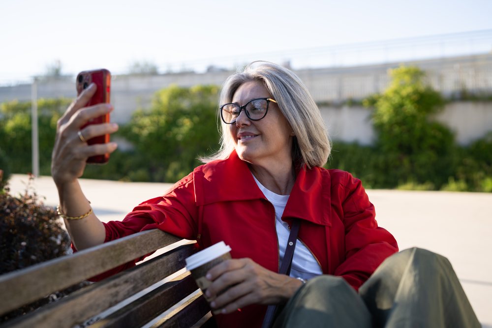 Middle-aged woman with gray hair sitting on a wooden bench outdoors holding a coffee cup and taking a selfie with a smartphone, Spain.