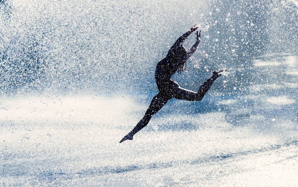 Woman performing a jumping acrobatic yoga pose in water spray with a silhouette effect, partner, acrobatics, USA.