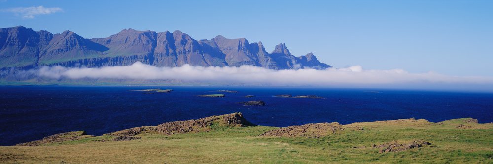 Panoramic view of a mountain range and ocean coastline under a clear blue sky with grassy rocky shoreline.