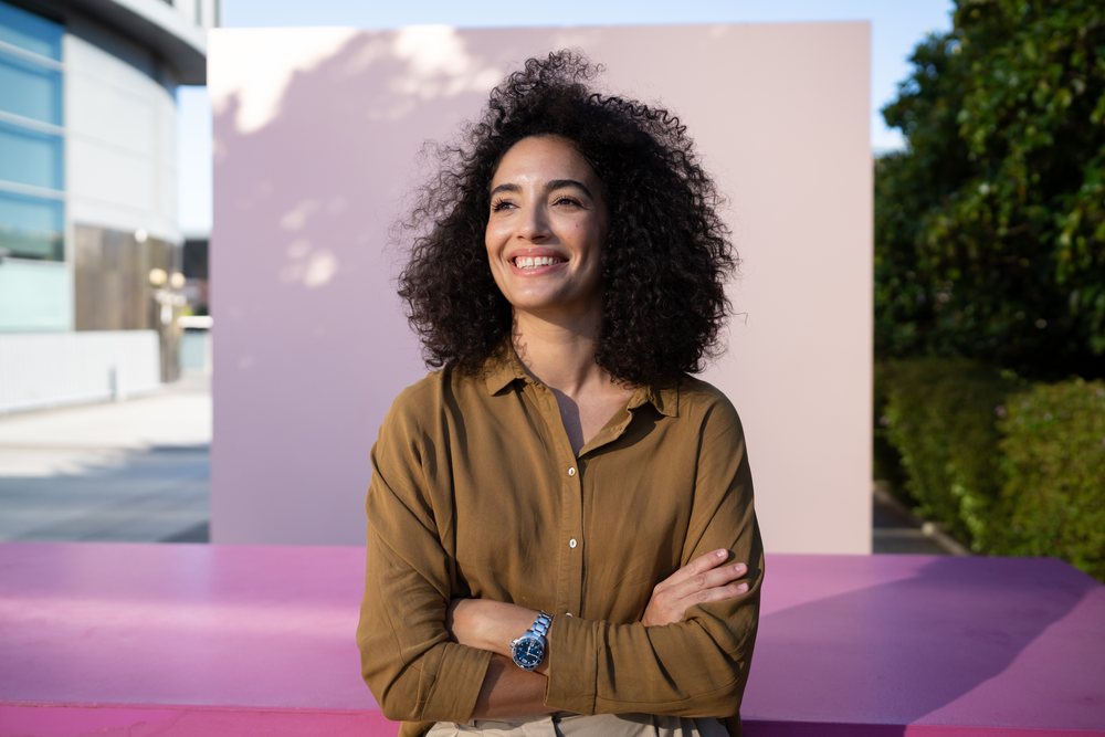 Woman with curly hair and brown shirt smiling outdoors in an urban setting with greenery and pink surface under natural sunlight, Madrid, Spain.