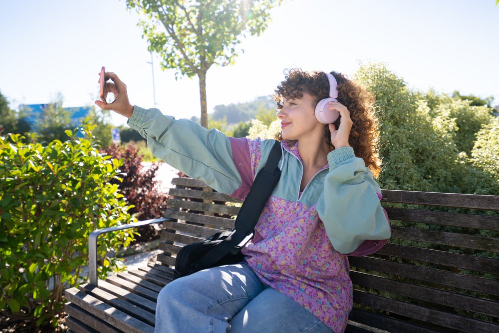 Woman with curly hair wearing pink headphones and a patterned jacket takes a selfie on a wooden bench outdoors, Spain.