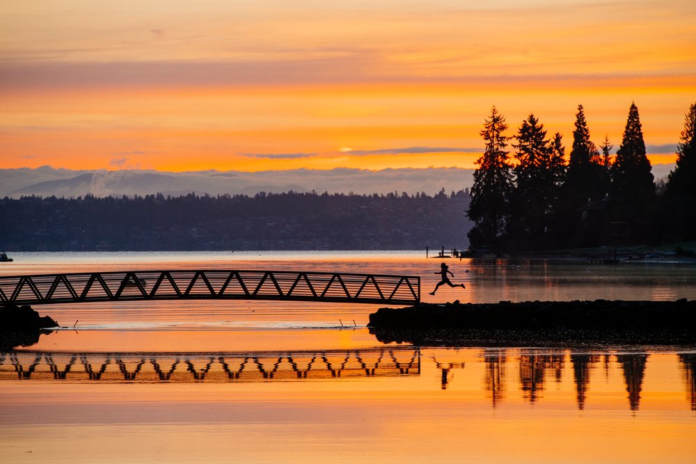 Silhouette of a person jumping near a pier at sunset over calm water on Bainbridge Island with trees along the shoreline, WA, USA.
