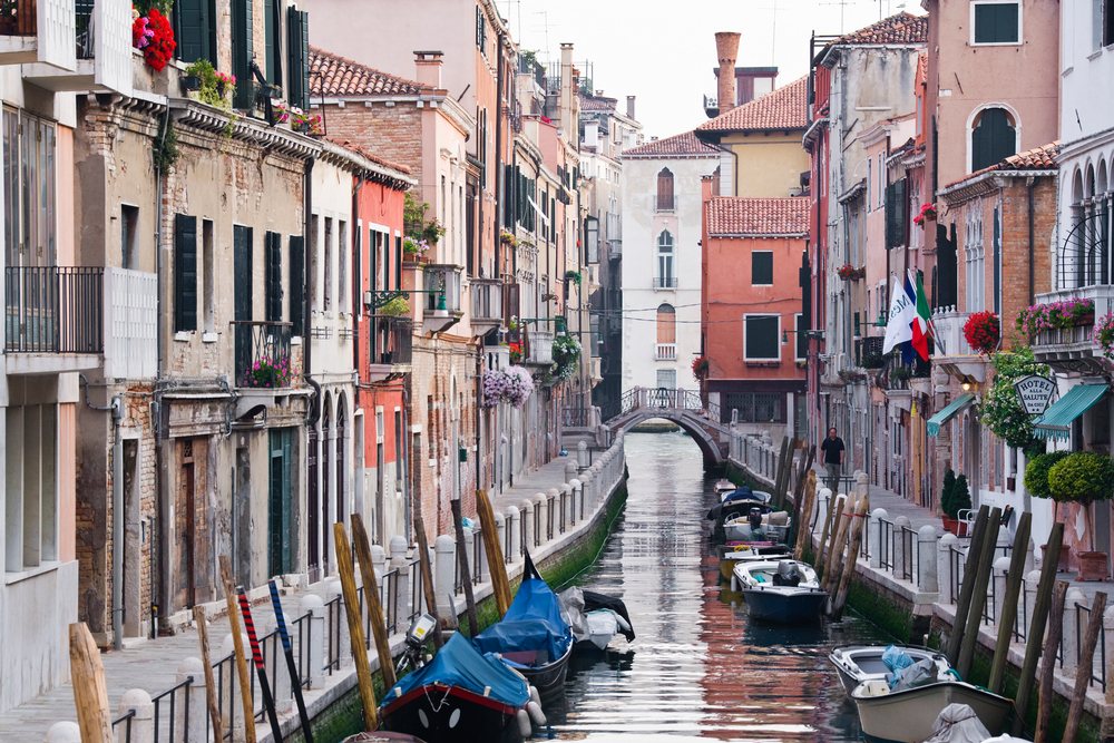 Venice canal with moored boats along narrow waterway flanked by historic buildings and an arched stone bridge, Italy.