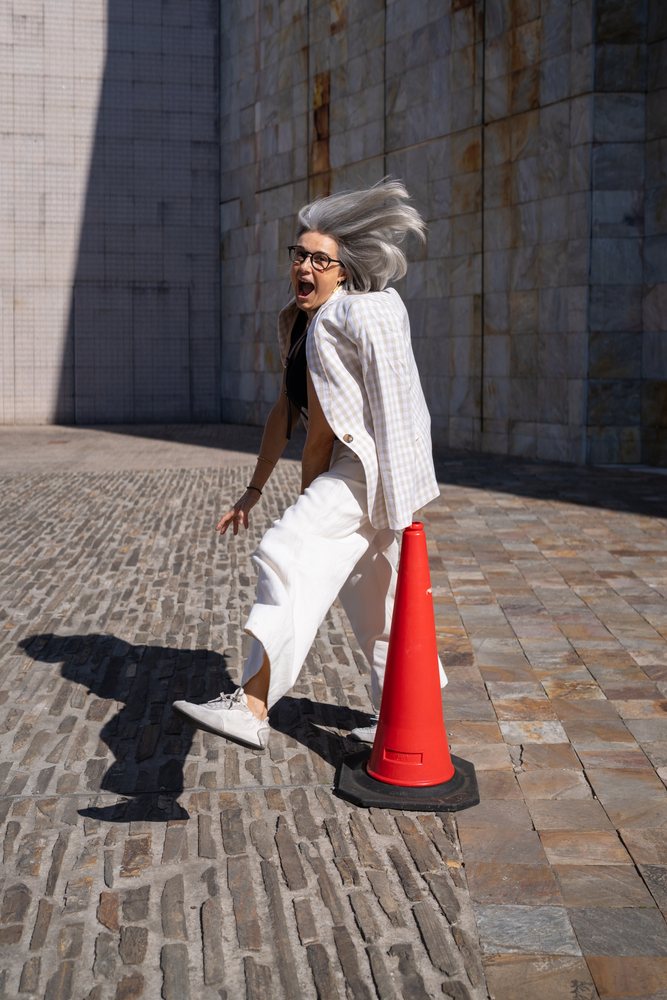 Woman with grey hair and glasses walking near a red traffic cone on a paved plaza outside a stone building in sunlight, businesswomen, Spain.