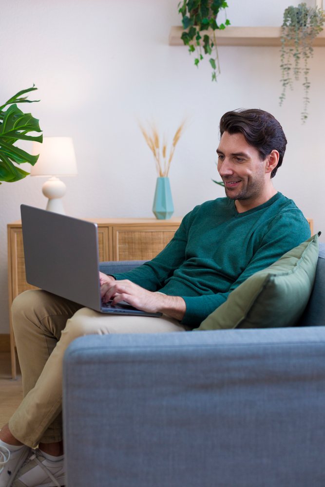 Man sitting on a sofa using a laptop in a modern home living room with plants and wooden furniture, Technology, young adult.