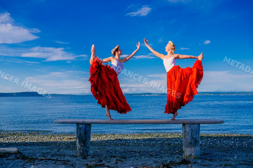 Two white women practice partner yoga acrobatics on a bench by the sea