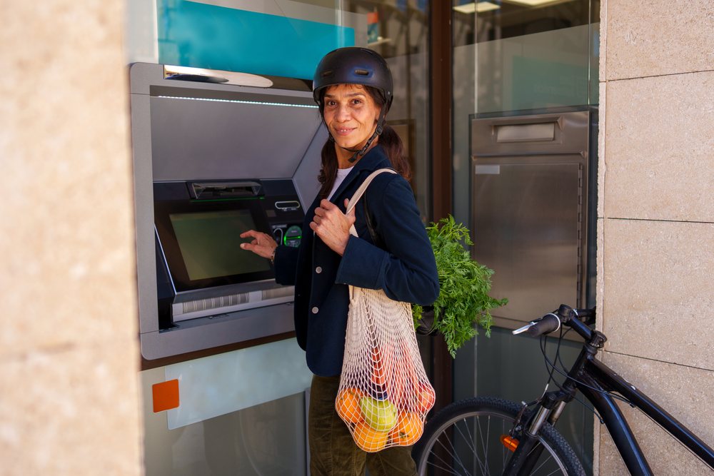 Woman wearing a helmet uses an ATM outdoors with a reusable net bag holding carrots, oranges, and an apple beside her bicycle, Málaga.