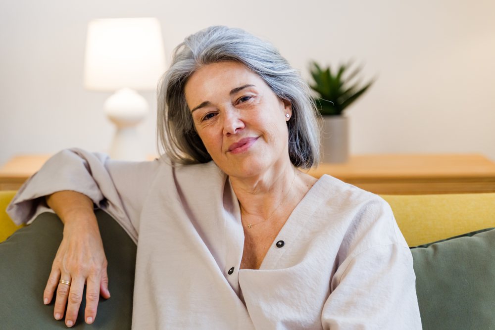 Older woman with gray hair sitting relaxed on a yellow couch in a modern living room with a lamp and houseplant behind her, Spain.