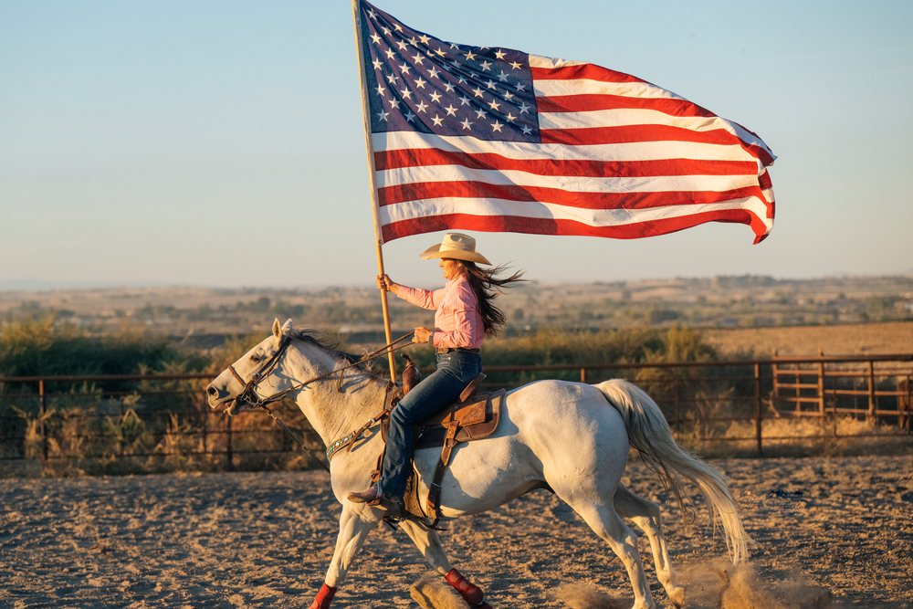 Woman wearing a cowboy hat, jeans, and boots rides a white horse holding an American flag in a desert ranch environment, Southwest USA.