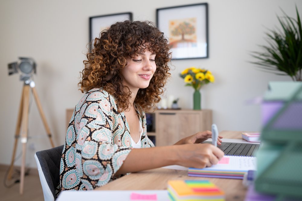 Woman with curly hair writing in a notebook at a home office desk with a computer keyboard and a vase of sunflowers, Spain.