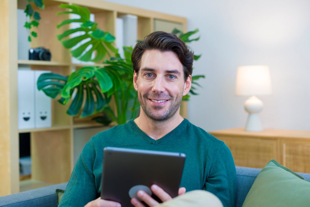 Man smiles while seated on a couch holding a tablet in a cozy home living room with plants and a lamp, Technology, young adult.