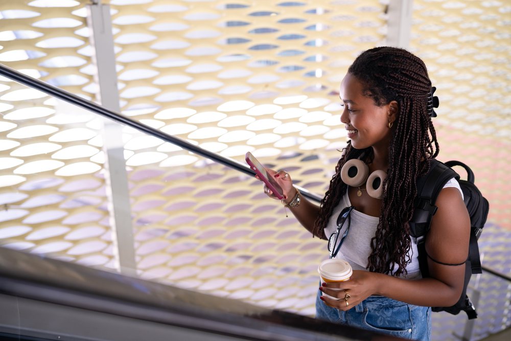 A young woman holding a coffee cup and using her smartphone while riding an escalator inside a modern building, remote work, travel, Santiago De Compostela, Spain.