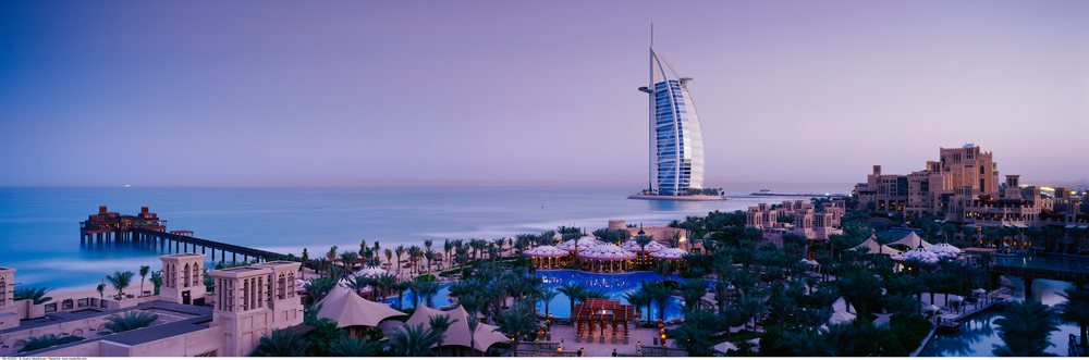 The Burj Al Arab hotel stands prominently on the Dubai coastline at dusk with nearby resorts and palm trees along the beachfront.