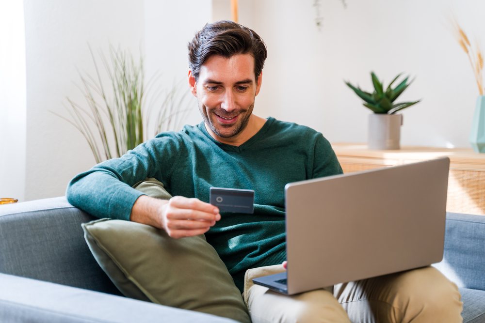 Man sitting on a sofa holding a credit card and using a laptop in a bright home interior with indoor plants and cushions, Spain.