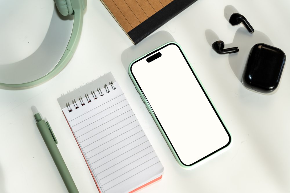 Smartphone with screen white and black wireless earbuds alongside spiral notepad, pen, and mirror on a white desk, mock up, white screen, Spain.