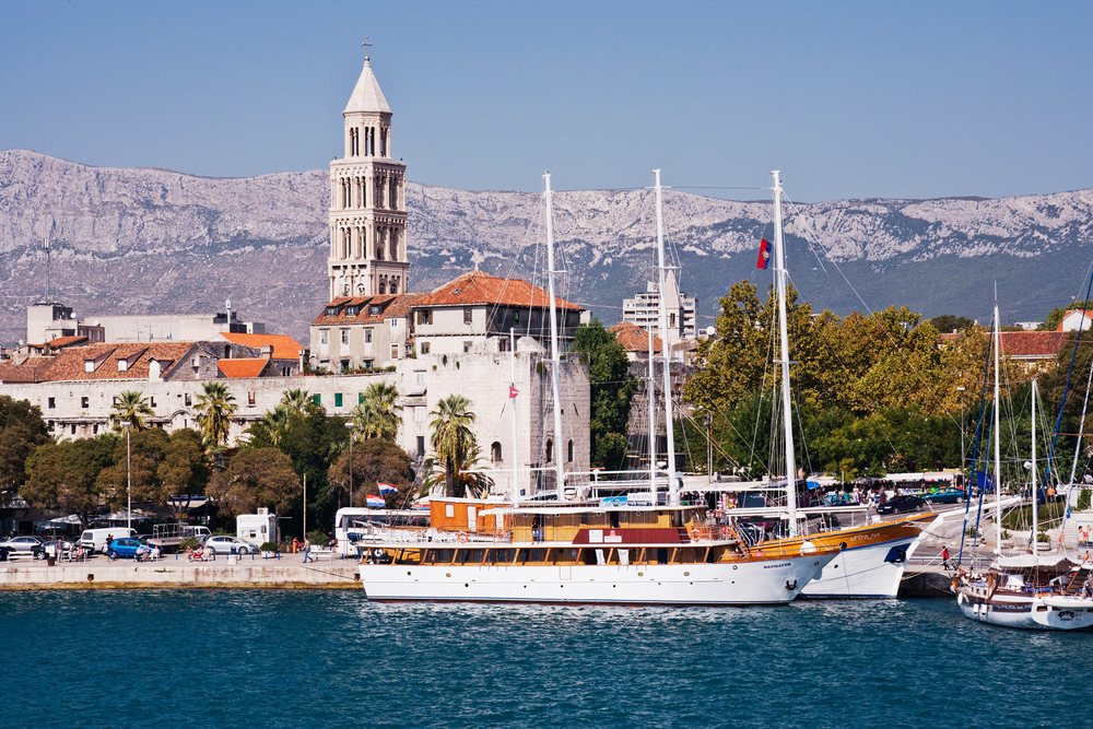 Harbor, Croatia with boats docked near historic buildings and a tall stone tower, mountains in background.