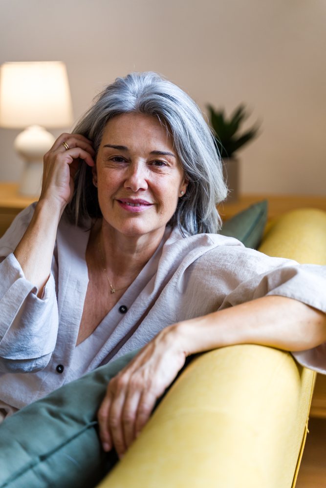 Middle-aged woman with gray hair smiles while relaxing on a yellow sofa in a living room with a lamp and indoor plant, Spain.