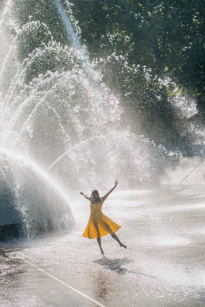 Woman in a yellow dress dances joyfully in a large public fountain spraying water on a sunny day outdoors, USA.