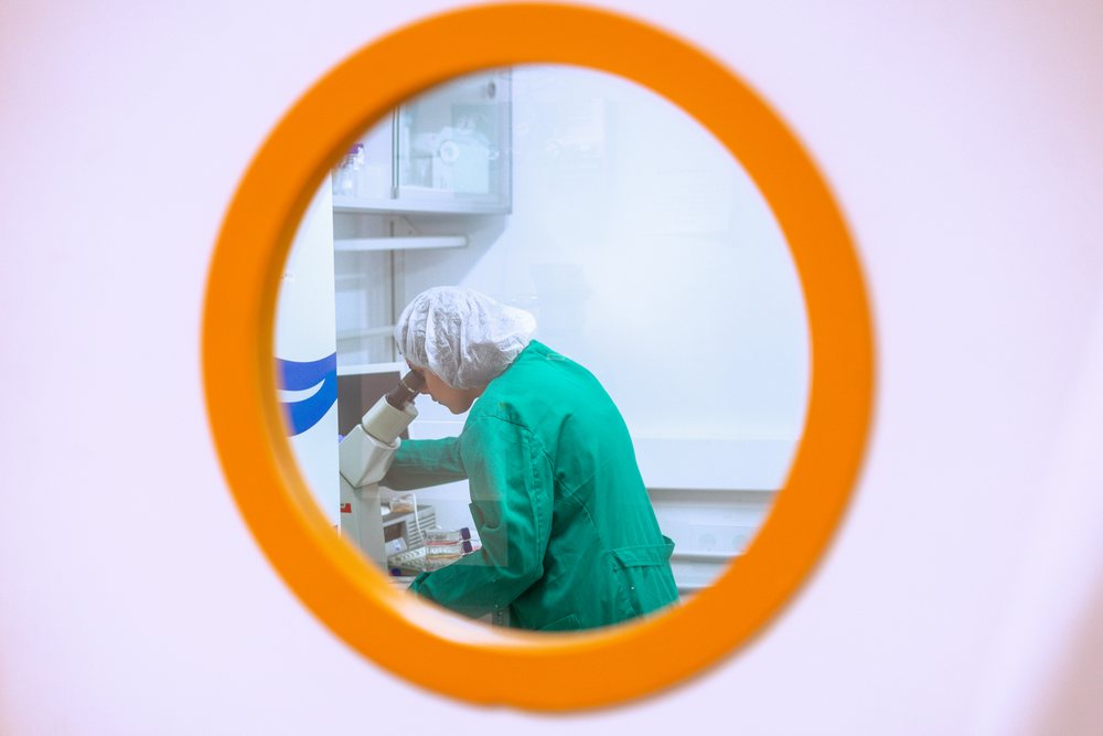 Scientist in green lab coat and hair net looking into microscope inside a laboratory, research, biology, Seville, Andalucía.