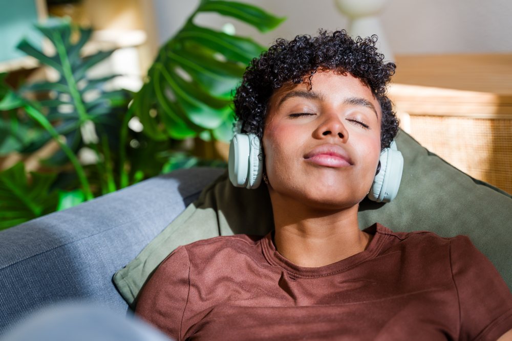 Woman with curly hair wearing brown shirt and white headphones relaxes on a couch supported by a green pillow near a monstera plant indoors, Spain.