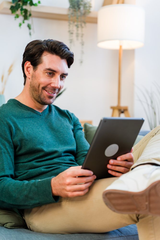Man sitting on a couch indoors in a modern living room holding and using a tablet with a smile, Spain.