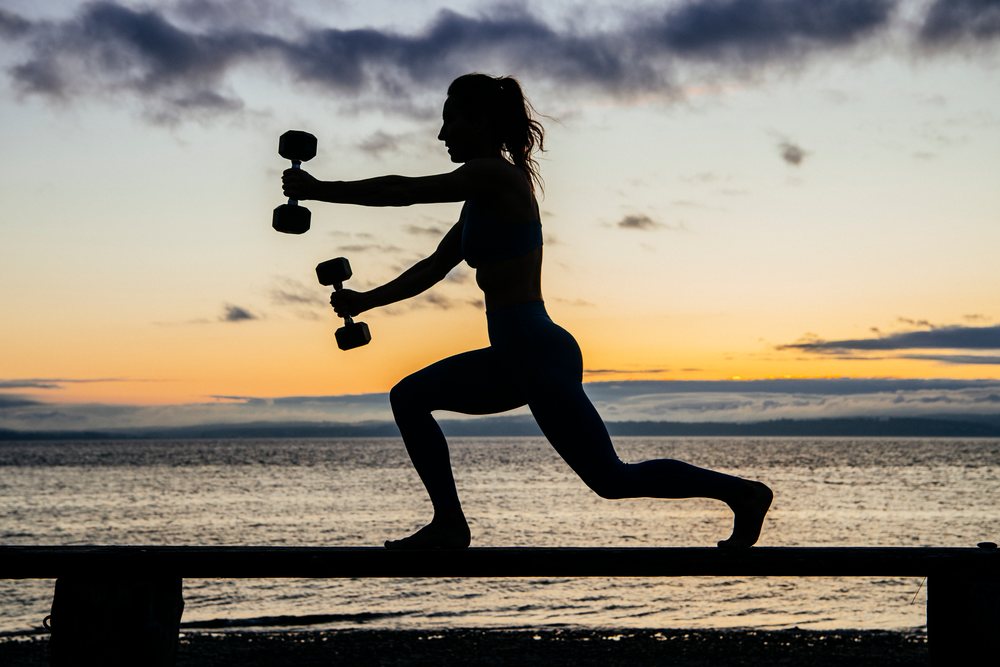 Woman performs a lunge exercise holding dumbbells on a bench by the sea at sunset, WA, USA.