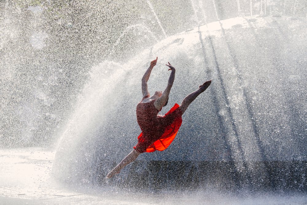 Female dancer in a red dress leaps gracefully through curved water spray from an outdoor fountain, USA.