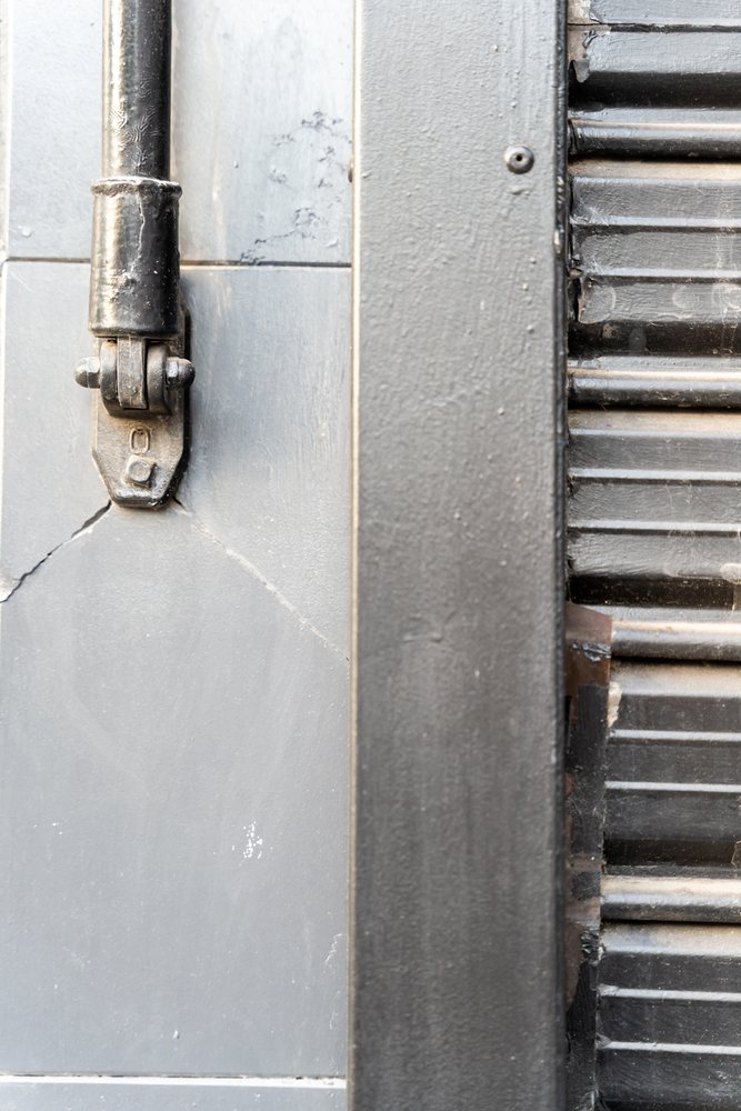 Close-up of a black metal hinge mounted vertically on a gray textured wall next to a slatted black metal shutter, texture, abstract texture, background, wallpaper, Spain.