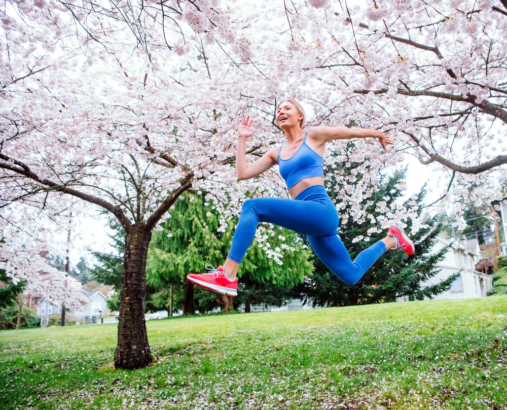 Woman jumps in athletic wear under blooming cherry blossom trees in an outdoor park during spring, USA.