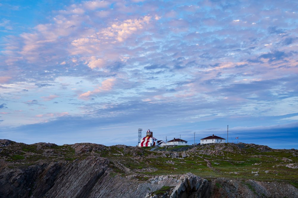 Cape Bonavista Lighthouse stands atop a rocky hill with multiple buildings and a Canada flag on a flagpole at Sunset, Landscape, Cityscape, Lifestyle, Newfoundland & Labrador.