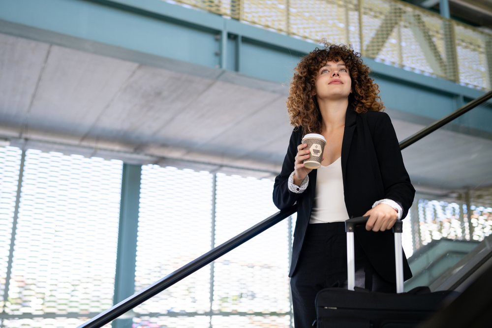 Woman holding coffee cup and pulling suitcase on escalator inside modern building, Spain.