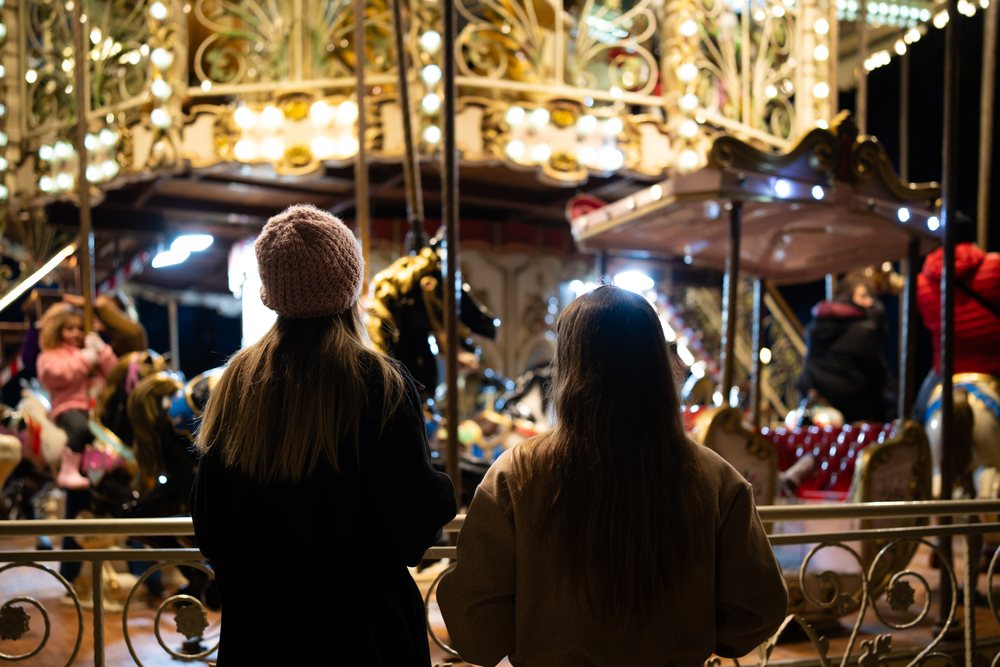Two women watch children riding a brightly lit carousel at a Christmas market in Madrid at night, Spain.