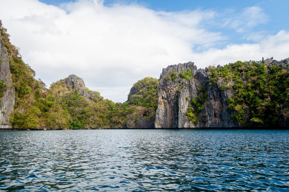 Limestone cliffs with tropical vegetation rise above calm blue water under a partly cloudy sky, Philippines.
