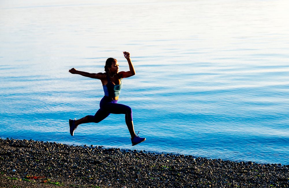 Woman in athletic wear is jumping on a pebbled shoreline beside a calm water surface, yoga, acrobatics, USA.
