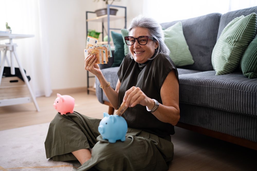 Woman with gray hair and glasses smiles while inserting euro banknotes into a blue piggy bank on her lap in a living room setting, Spain.