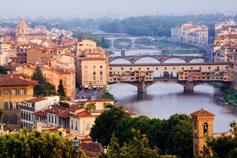 View of the Arno River flowing through Florence with the historic Ponte Vecchio bridge and dense urban buildings during daylight, Piazza Michelangelo, Italy.