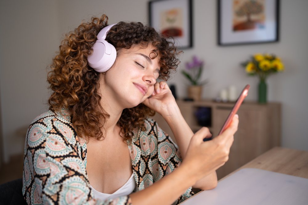 Woman with curly hair listens to music using headphones while holding a smartphone at home, Spain.