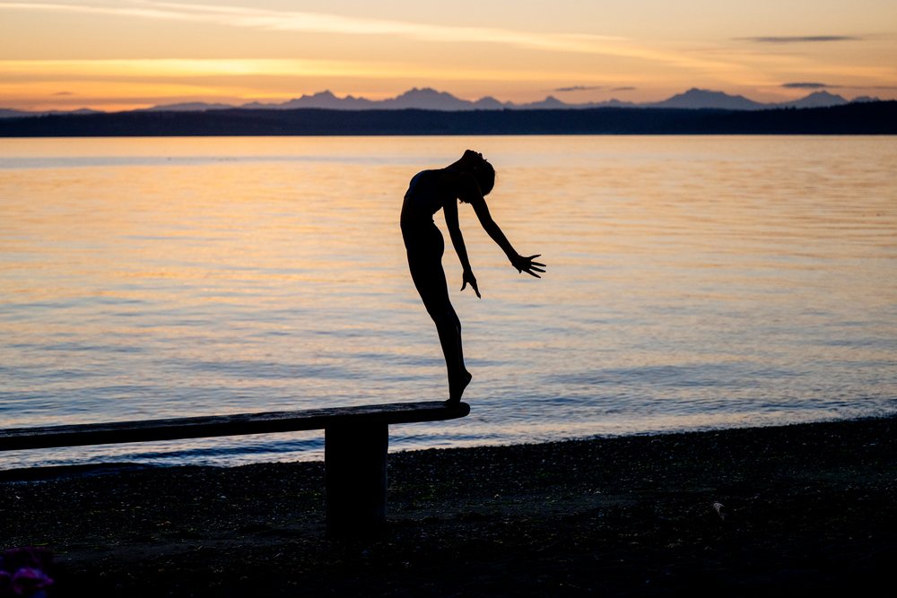 Woman practicing yoga acrobatics balancing on a bench by the lake at sunset, USA.