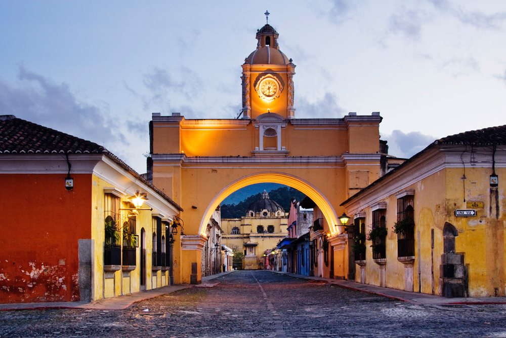 Santa Catarina Arch with illuminated clock tower and colonial buildings along cobblestone street at twilight, Antigua, Guatemala.