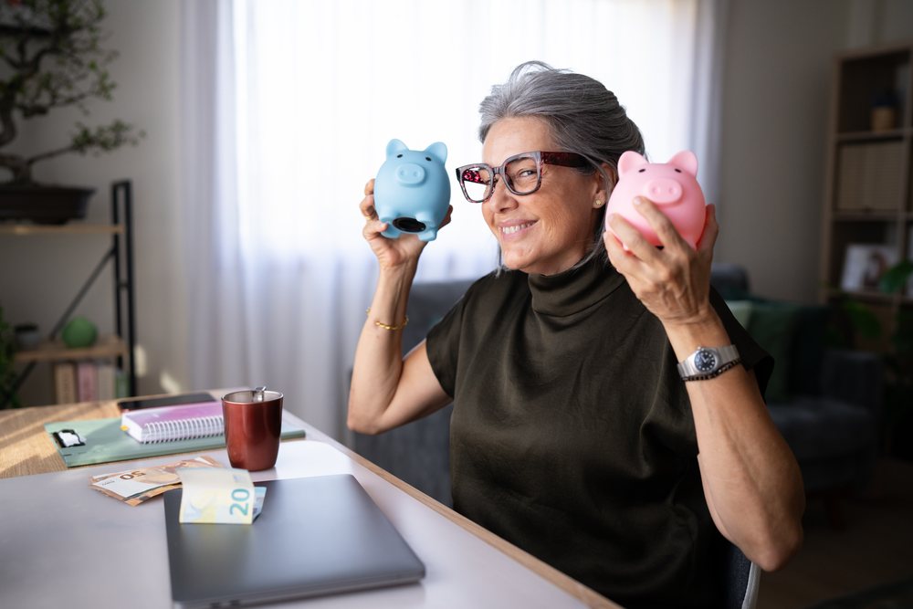 Senior woman in a home office holding blue and pink piggy banks with money and office supplies on her desk, Spain.